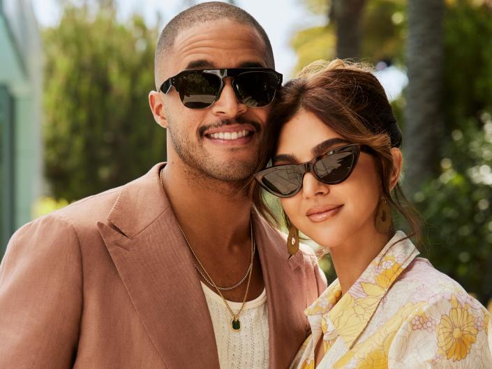 Ray Jimenez is a man in a brown jacket, white shirt and necklace, wearing sunglasses and posing next to his wife Eilyn Jimenez who is wearing a yellow and white floral jacket and sunglasses