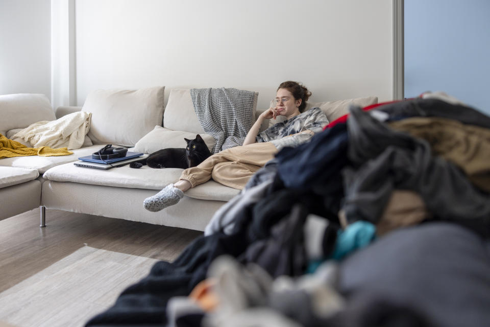 Person lounging on a sofa with a black cat, a stack of clothing on the floor. Mood is relaxed and casual