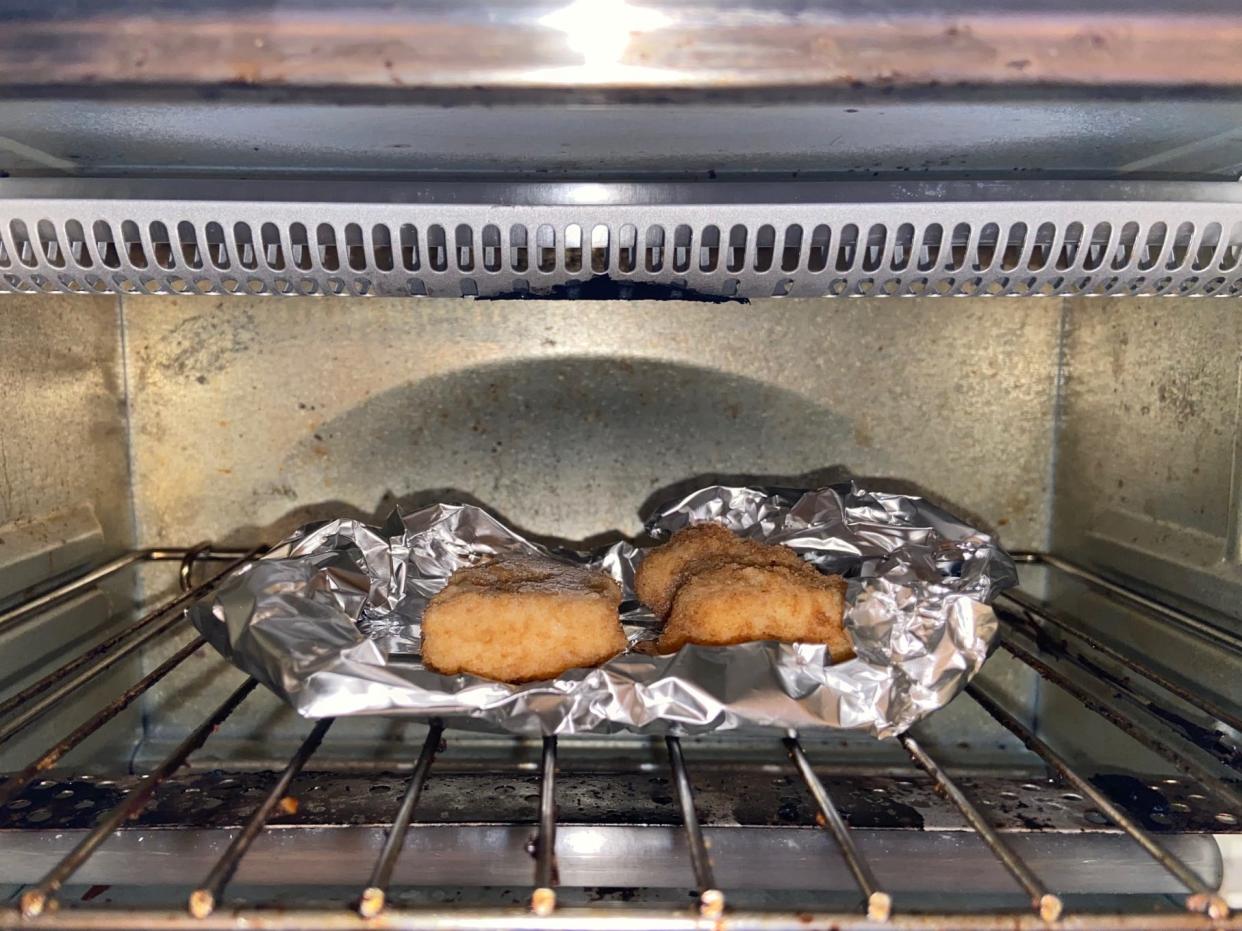 Two chicken nuggets in an aluminum foil faux bowl in the toaster oven on a metal grate.