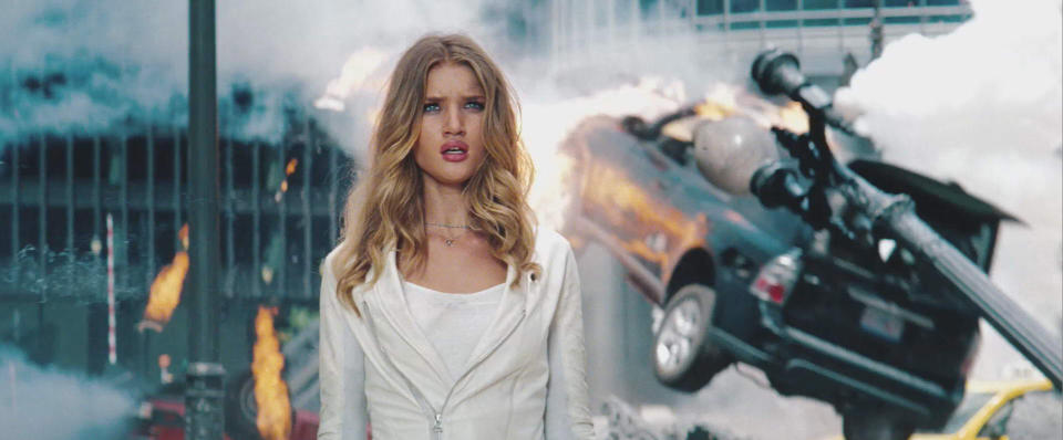 Rosie Huntington-Whiteley in an intense action scene with a car crash and explosions in the background