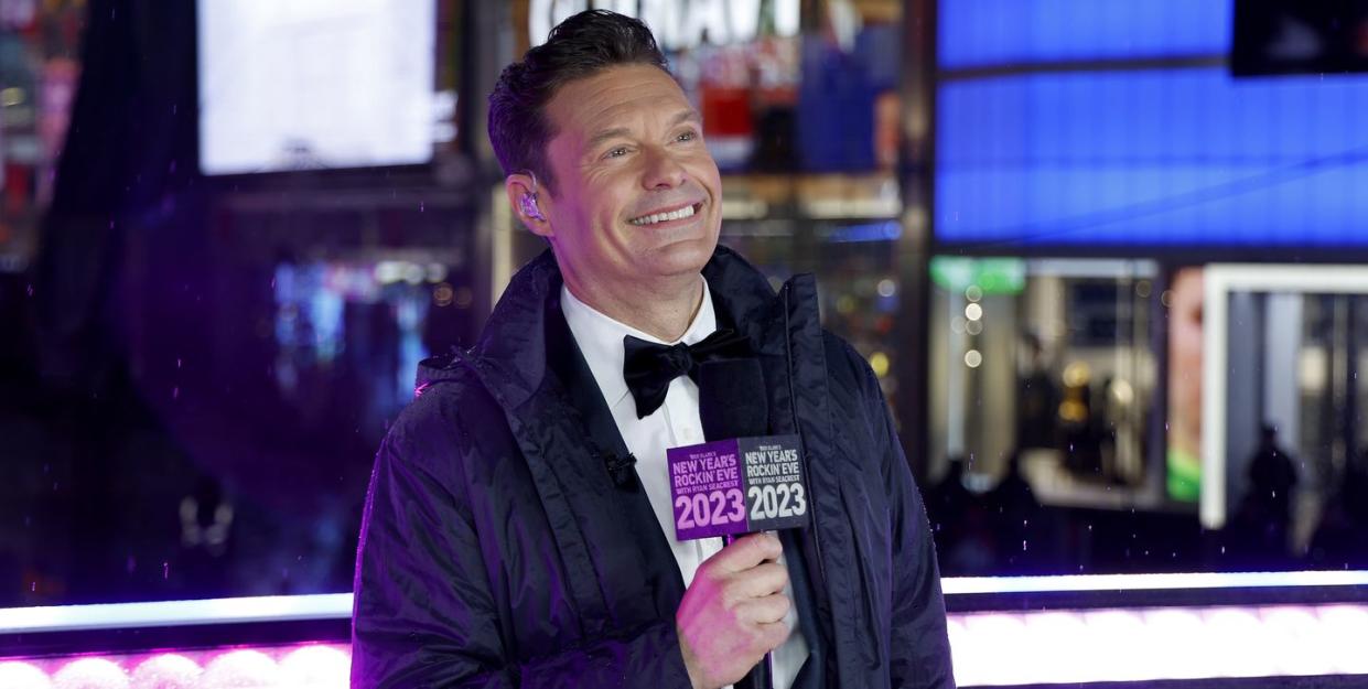 ryan seacrest wearing a tuxedo and jacket as he smiles off camera while holding a microphone