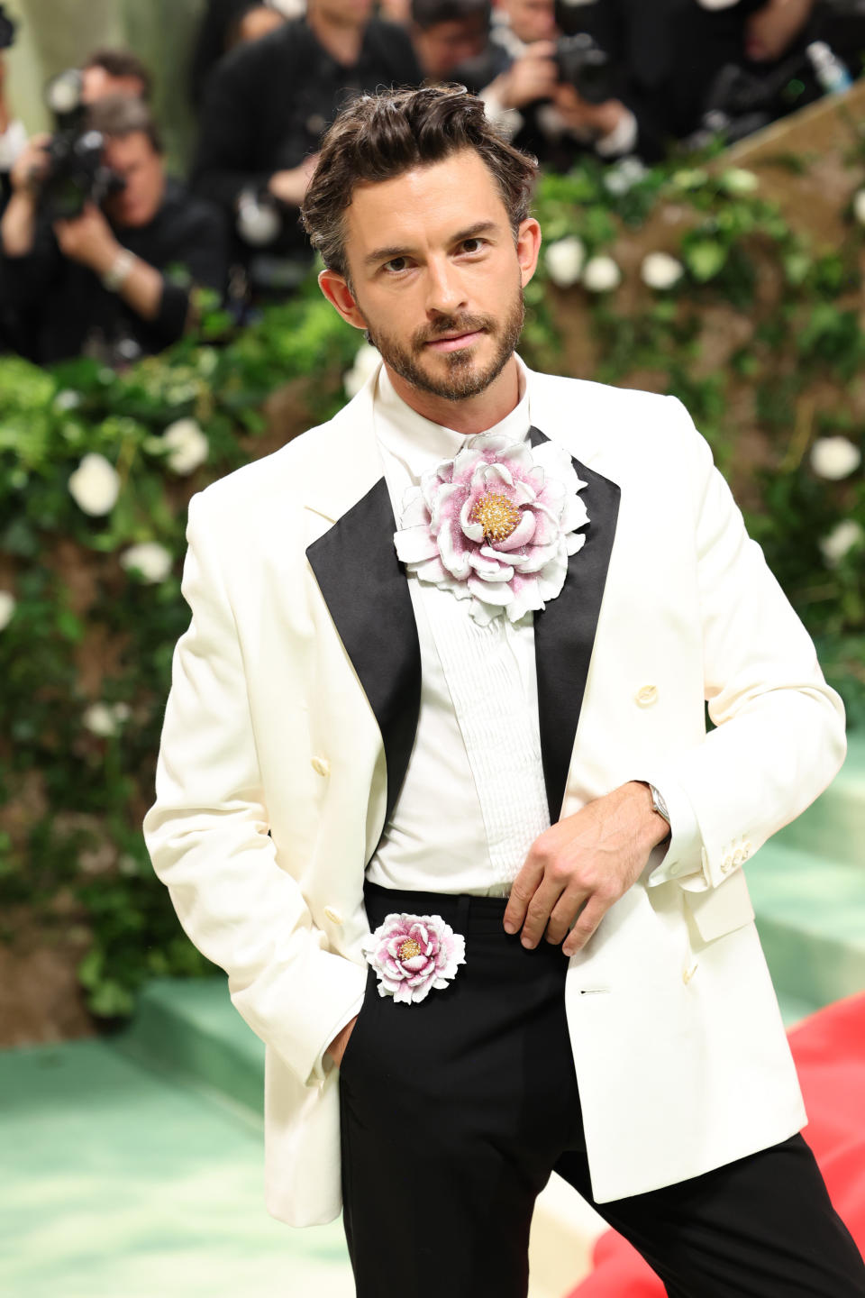 Jonathan Bailey in white tuxedo with large floral adornment on lapel, hands in pockets, posing at the Met Gala in 2024