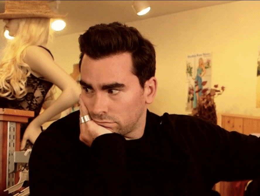 Dan Levy, wearing a black outfit, rests his head on his hand with a contemplative expression in a clothing store. Text: #SchittsCreek