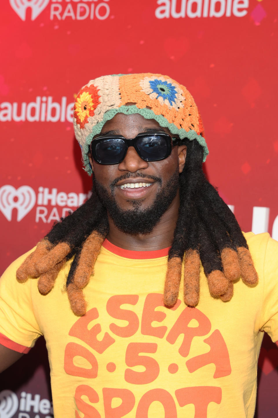 Man in crocheted hat and sunglasses on a red carpet, wearing a yellow "Desert 5 Spot" shirt