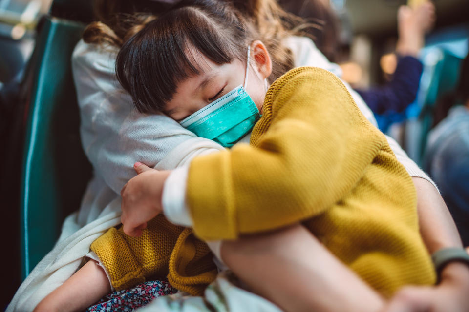 Lovely little girl with medical face mask sleeping soundly in mother’s arms while they are riding on bus.