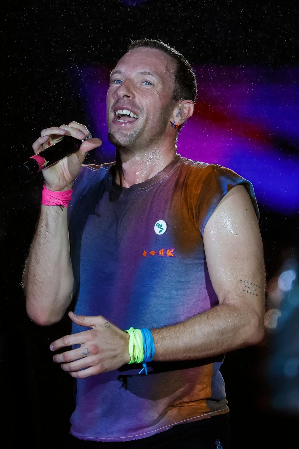 Chris Martin performing on stage, wearing a sleeveless top with a smiley badge, wristbands, and singing into a microphone