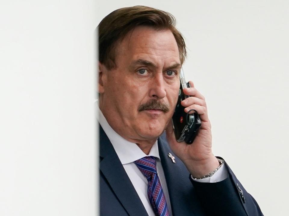 Mike Lindell leaning on a white wall while taking a phone call and wearing a suit with a patterned tie.