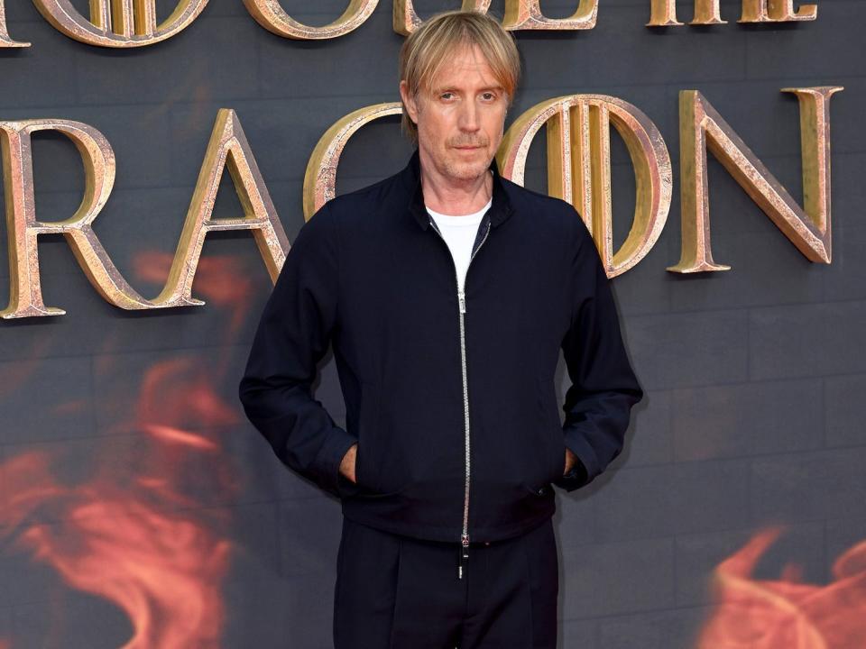 A man with blonde hair wearing a dark jacket and pants with his hands in his pocket.