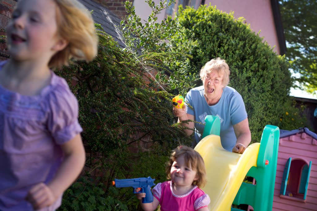 A senior citizen plays with two children outside that she's babysitting. They are chasing each other with water guns.