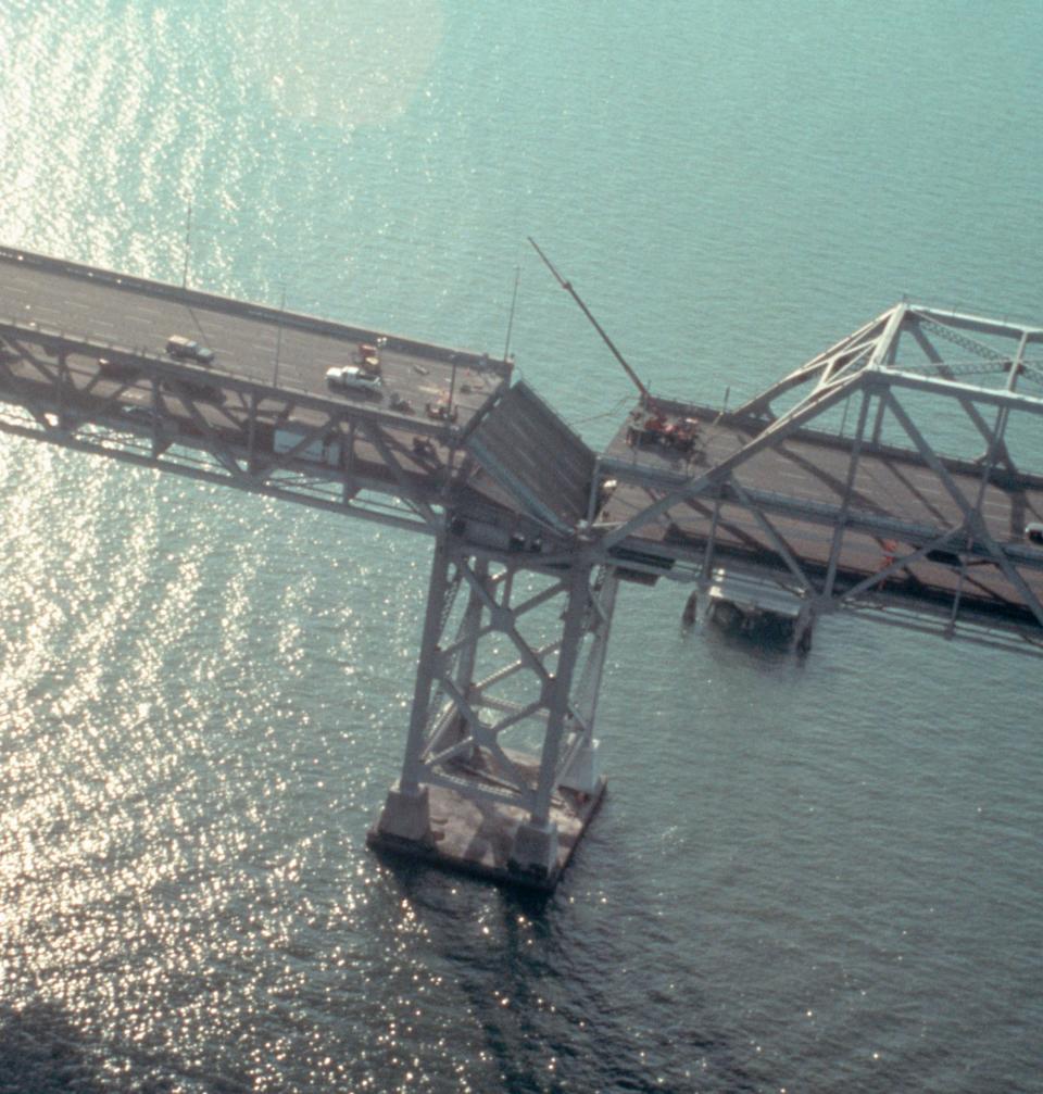 Partial collapse of a bridge over water, with a missing segment in the middle where vehicles are halted. The image shows structural damage and emergency activity