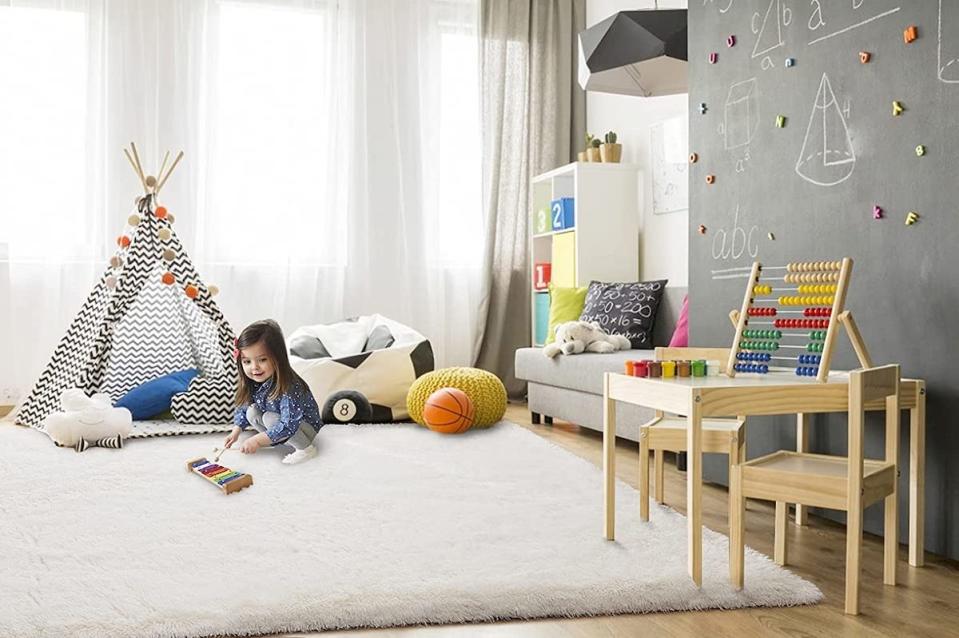 Young girl playing on a white furry rug in a playroom with a chalkboard wall, tent, abacus, and toys.