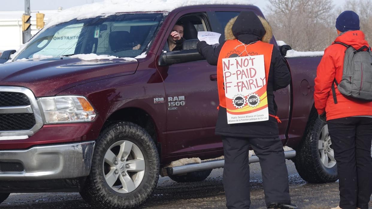 A picket hands out information to a motorist outside the Royal Military College in Kingston, Ont., on Jan. 15, 2024. Some 500 civilian staff members at bases in Ontario and Quebec who walked off the job are calling for a national pay grid.  (Dan Taekema/CBC - image credit)