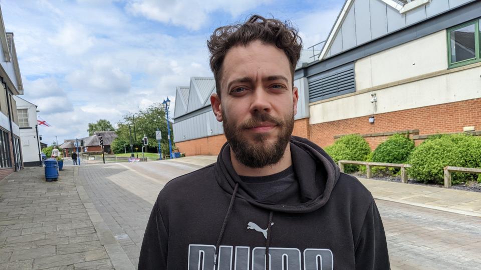 Daniel Newton wears a hoodie and looks at the camera as he stands on a street