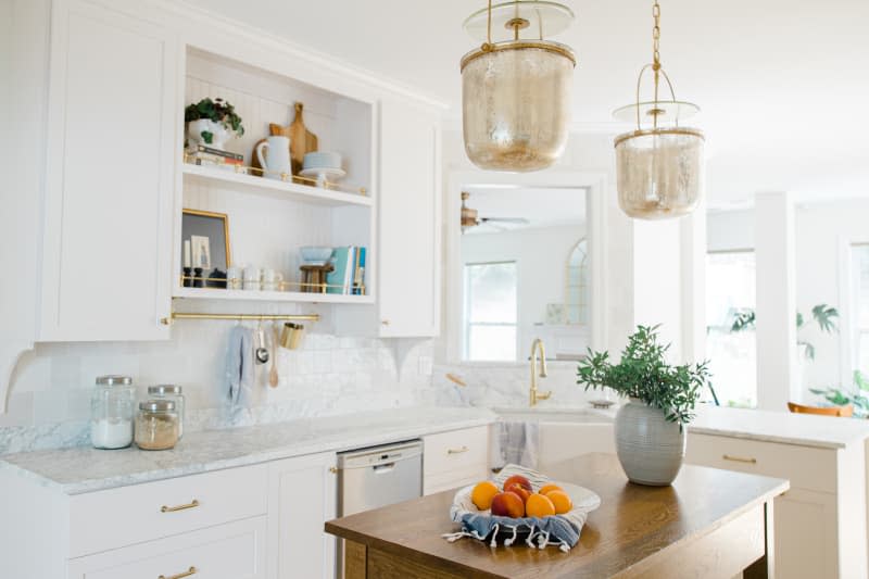 Renovated white kitchen features marble counters, tile backsplash, brass accents and pendant lamps.