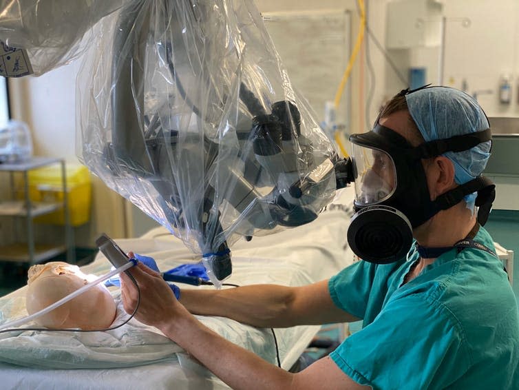 A surgeon in scrubs and a hairnet wears a respirator mask and looks down a microscope while simulating an operation on a skull.