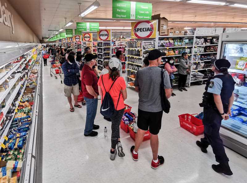 A police officer watches on at people queueing for a delivery of toilet paper, paper towel and pasta at Coles Supermarket, Epping in Sydney.