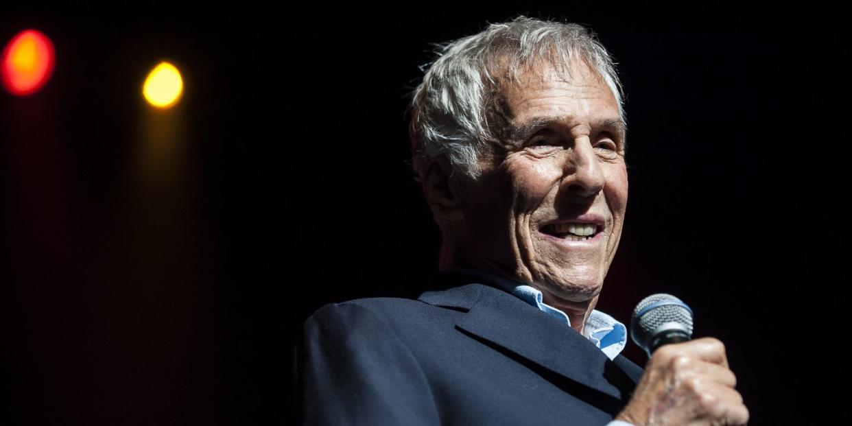 burt bacharach smiling and standing on a stage, holding a microphone and wearing a suit jacket