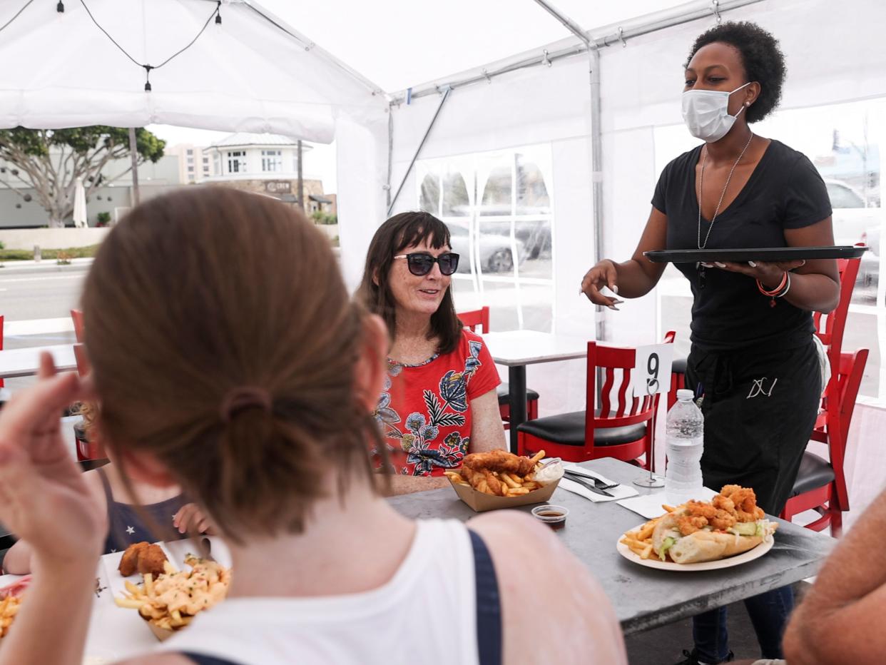 A young black woman serves food to customers sitting inside a tent. She is wearing a mask