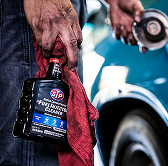 A man's hand is holding a spray bottle of STP fuel injection cleaner his another hand is shown holding a car's gas cap open.