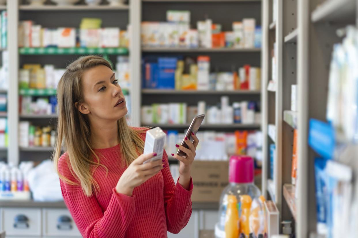 Profile shot of beautiful young woman shopping at pharmacy. Woman holding medication container while talking on cell phone.  Woman holding medication box and dialing on cell phone