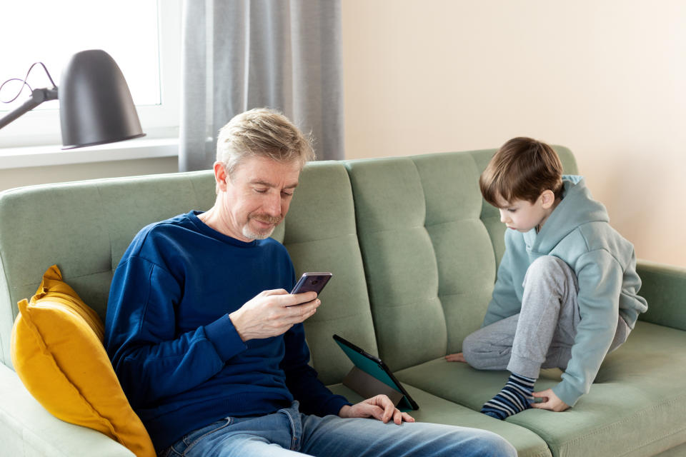 A man with a smartphone sits on a couch next to a young boy who is looking at a tablet