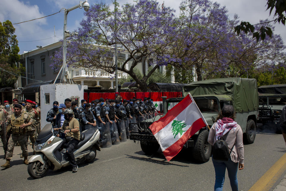 FILE - In this May 7, 2020 file photo, an anti-government protester walks by security forces holding a Lebanese flag during ongoing protests, outside the military court in Beirut, Lebanon. A year after anti-government protests roiled Lebanon, dozens of protesters are being tried before military courts that human rights lawyers say grossly violate due process and fail to investigate allegations of torture and abuse. (AP Photo/Hassan Ammar, File)