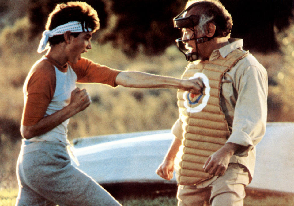 A scene from 'The Karate Kid' with Daniel Larusso practicing a punch on Mr. Miyagi wearing protective body gear