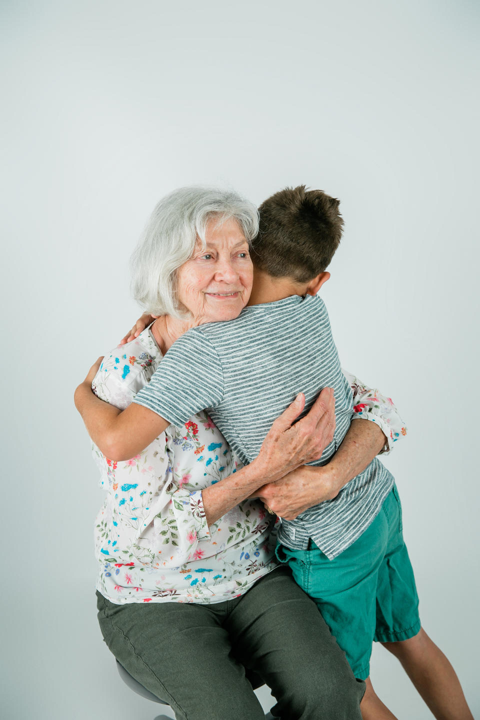 An elderly woman with gray hair, wearing a floral top, embraces a young boy in a striped shirt and green shorts.