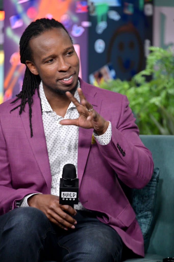 ibram x kendi sits on a couch and holds his hand in front of his chest as the other hand holds a microphone at his lap, he smiles and wears a magenta suit jacket, patterned collared shirt and jeans