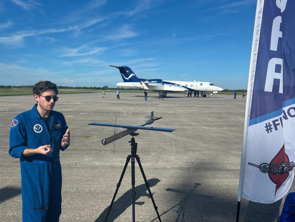 an aviator in a blue flight suit stands next to a small drone with a large white jet in the background