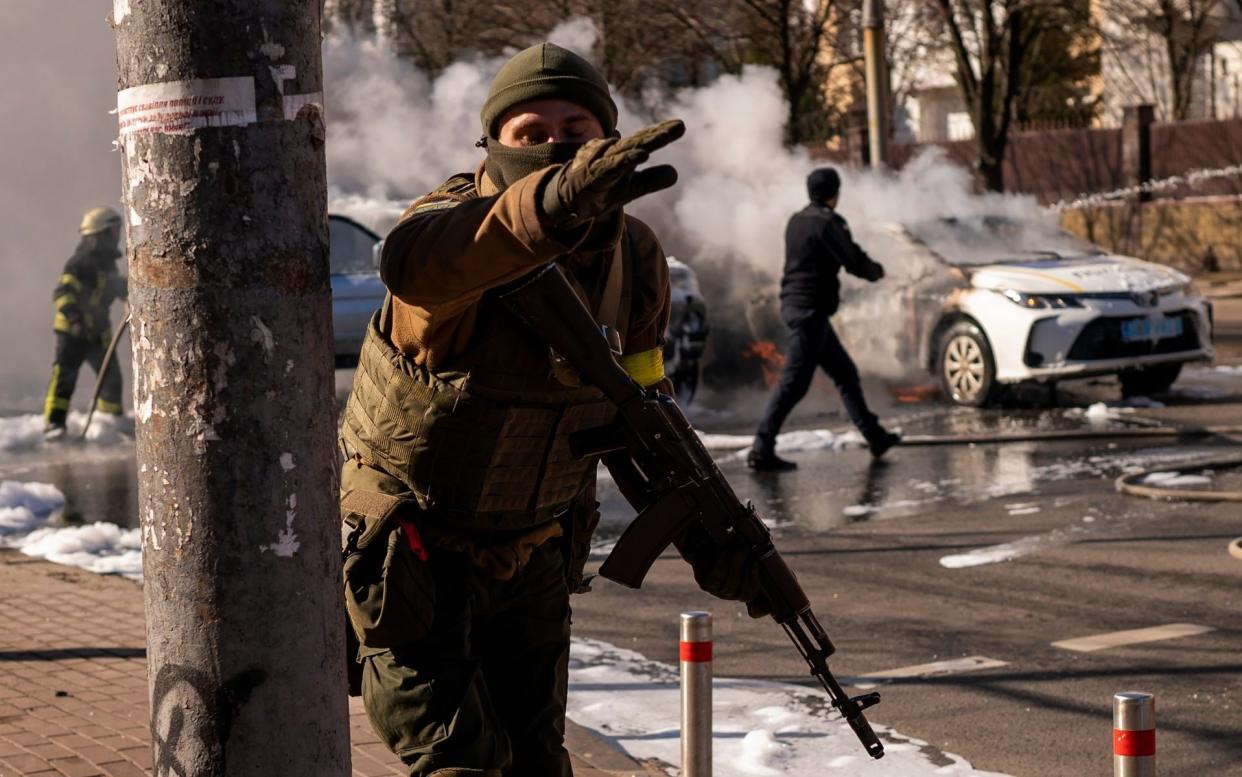 Ukrainian soldiers take positions outside a military facility as two cars burn in a street in Kyiv - Emilio Morenatti/AP
