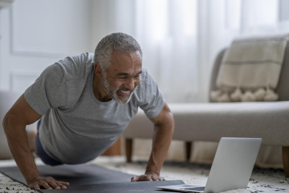 A senior gentleman of African decent, lays out on a yoga mat in his living room as he exercises along with a virtual class.  He is dressed comfortably in athletic wear and has his laptop out in front of him as he follows along with the instructor and does push-ups.