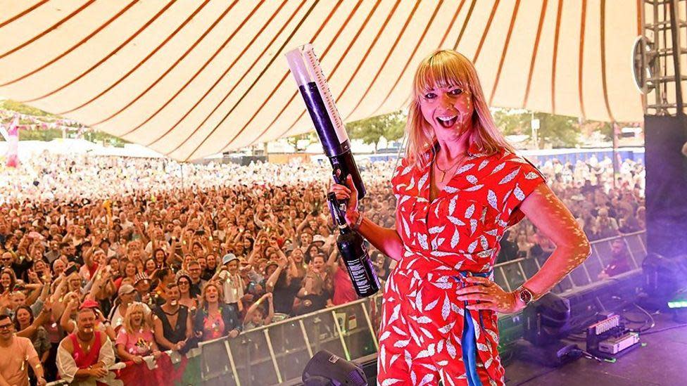 Sara Cox standing with her back to a festival crowd, grinning and holding a confetti cannon