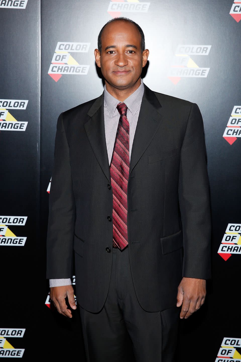 james rucker smiles at the camera while standing in a black suit, checkered shirt and red striped tie, he is in front of a black background with color of change logos