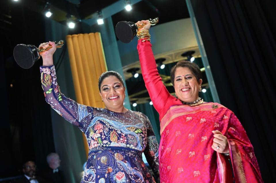 kartiki gonsalves and guneet monga, wearing a blue and red dress respectively, smile and hold academy awards in the air with their right arms
