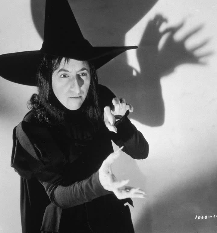 A person dressed as a witch poses dramatically with a pointed hat and a shadow on the wall
