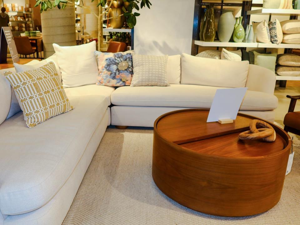 A white L-shaped couch with white pillows, pillows with geometric designs, and a floral pillow on it. A circular wooden table sits next to the sofa.