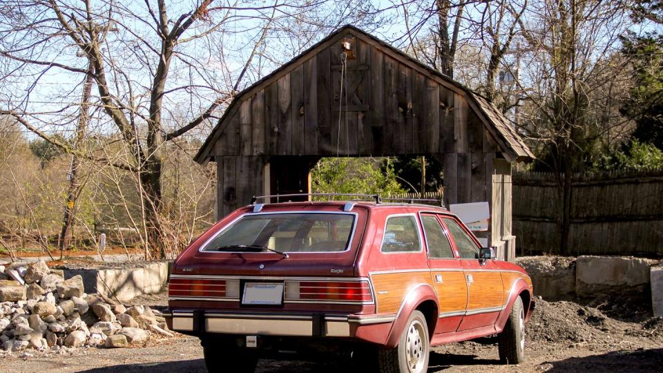 a red station wagon with woody siding sits in front of a barn and a small rock fence