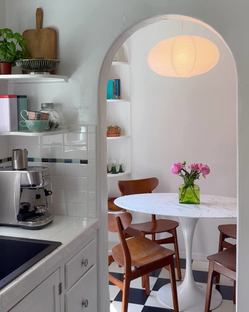 Paper mache oval lighting fixture, arched corner built in book cases, round white table with dark wood mid-century modern chairs, black and white checkered tile floor, arched door way from kitchen