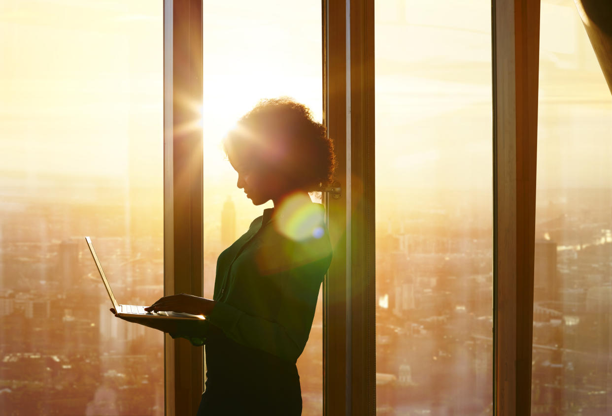 A woman working on a laptop computer while standing next to large windows above the city is seen in silhouette.