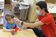 In this May 27, 2020 photo, Aaron Rainboth, a teacher at the Frederickson KinderCare daycare center in Tacoma, Wash., wears a mask as he takes the temperature of Benjamin Simpson, 4, after he complained of feeling hot following an outdoor play period, but found it to be normal. In a world weary of the coronavirus, many working parents with young children are now struggling with the decision on when or how they'll be comfortable returning to their child care providers. Frederickson KinderCare, which has been open throughout the pandemic to care for children of essential workers, removed carpets and spaced out tables and chairs as part of their measures to control the spread of the coronavirus. (AP Photo/Ted S. Warren)