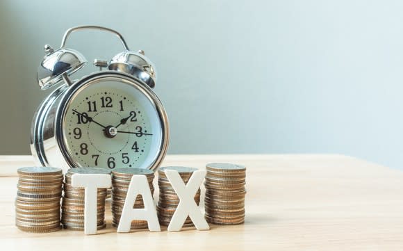 Silver clock with old-style alarm bells behind five piles of coins and letter magnets spelling tax, on a table.