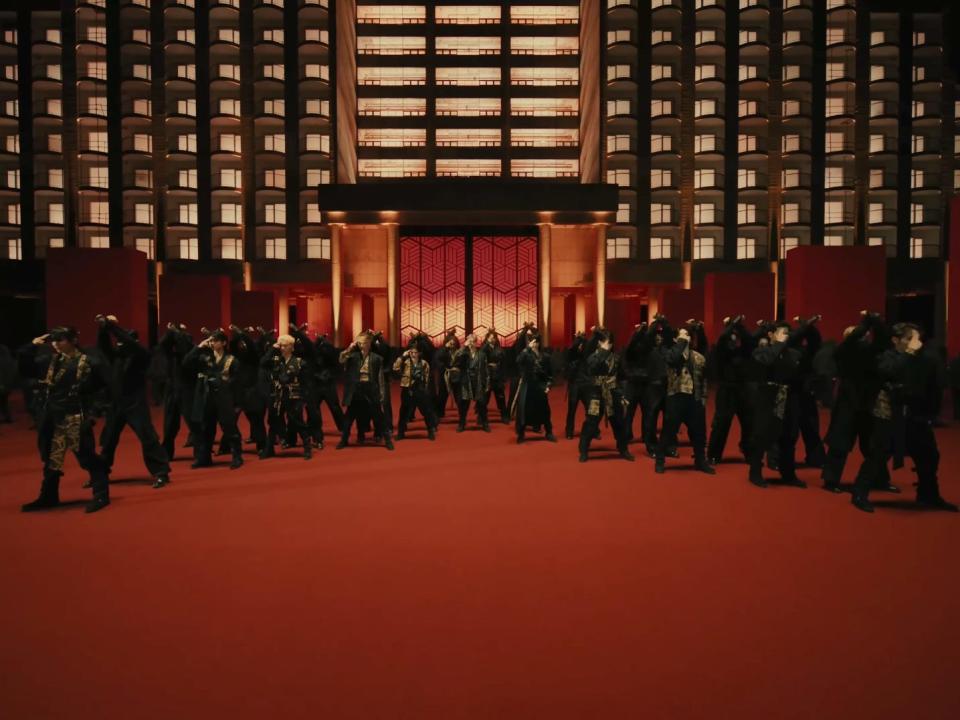 the thirteen members of seventeen dressed in black and gold outfits, with backup dancers in black clothing behind them. they're standing on a red surface with a golden palace behind them