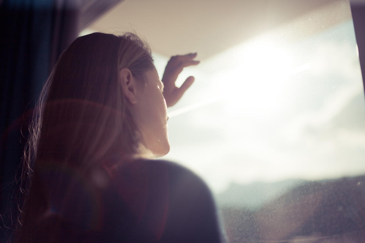 Blondhaired women looking out of window with back light in her face, Spain.