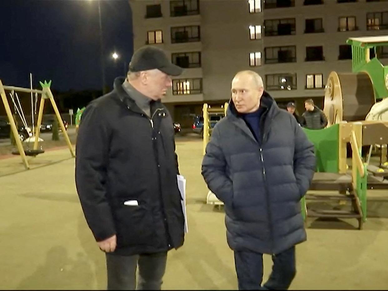 Russian President Vladimir Putin is wearing a black puffer jacket and pants and is accompanied by another man in a black puffer jacket and pants. They are both standing on a playground with a green slide and yellow swings. It is night time.