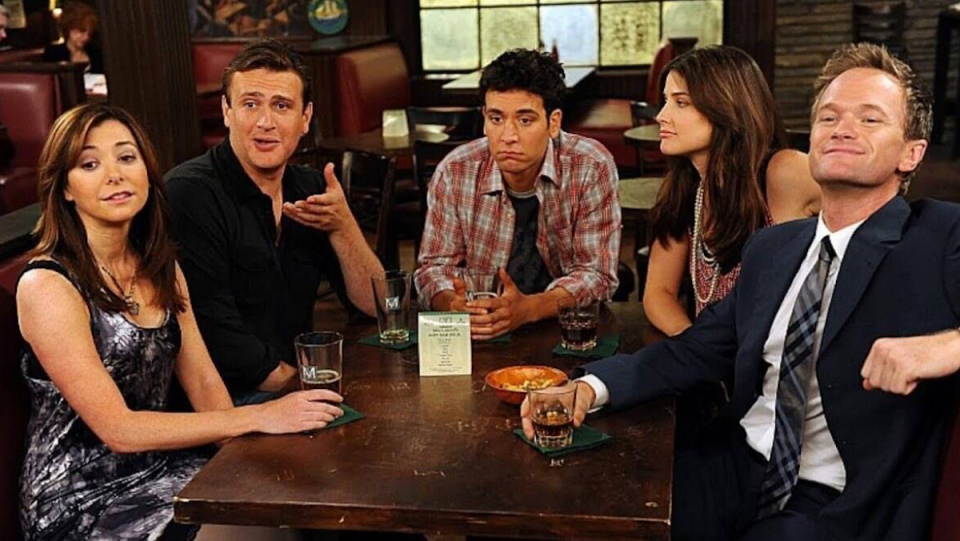 The cast of How I Met Your Mother sits around their bar booth.