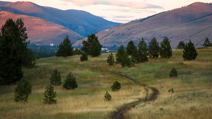 A trail winds through green hills with larger mountains in the background