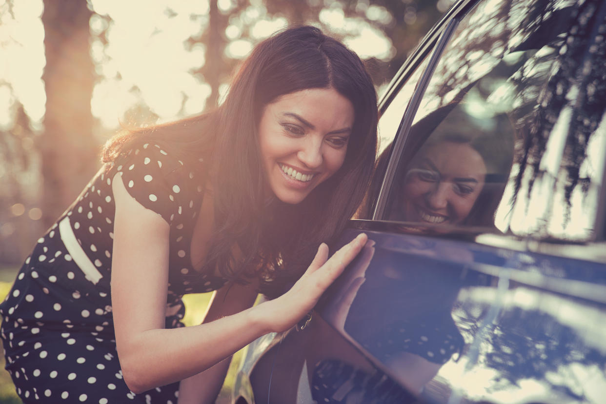 Excited young woman and her new car outdoors with sunlit forest in background.