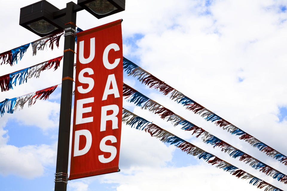 'Used Cars' sign over a vehicle dealership car lot. Red. Clouds in sky. 