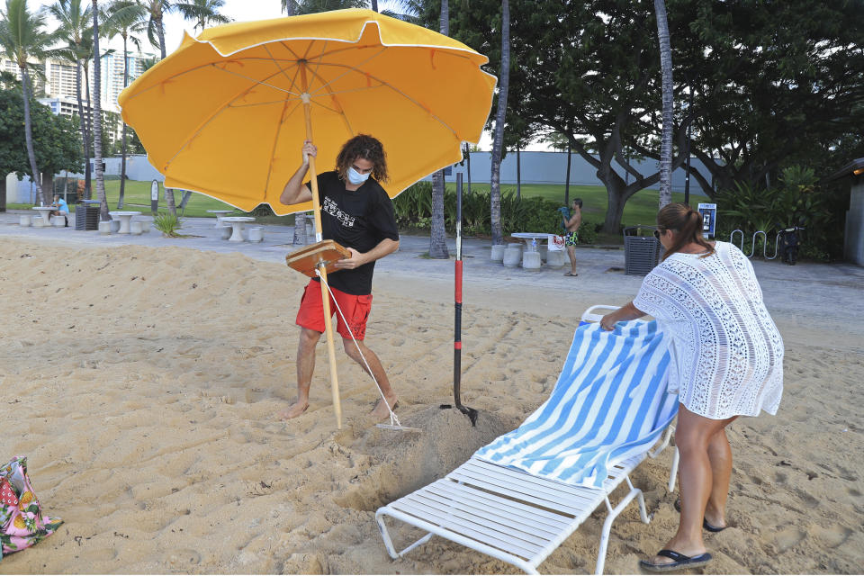 Mitch Kemp, left, a worker at a Waikiki Beach rental company, sets up an umbrella for visitor Leena Kilman on Waikiki Beach, Thursday, Oct. 15, 2020, in Honolulu. Kilman, who is visiting from New Hampshire, spent two weeks in mandatory quarantine and Thursday was her first day to visit the beach. The beach rental company opened for the first time Thursday since closing in March due to the COVID-19 pandemic. Kilman was their first rental customer since closing. (AP Photo/Marco Garcia)